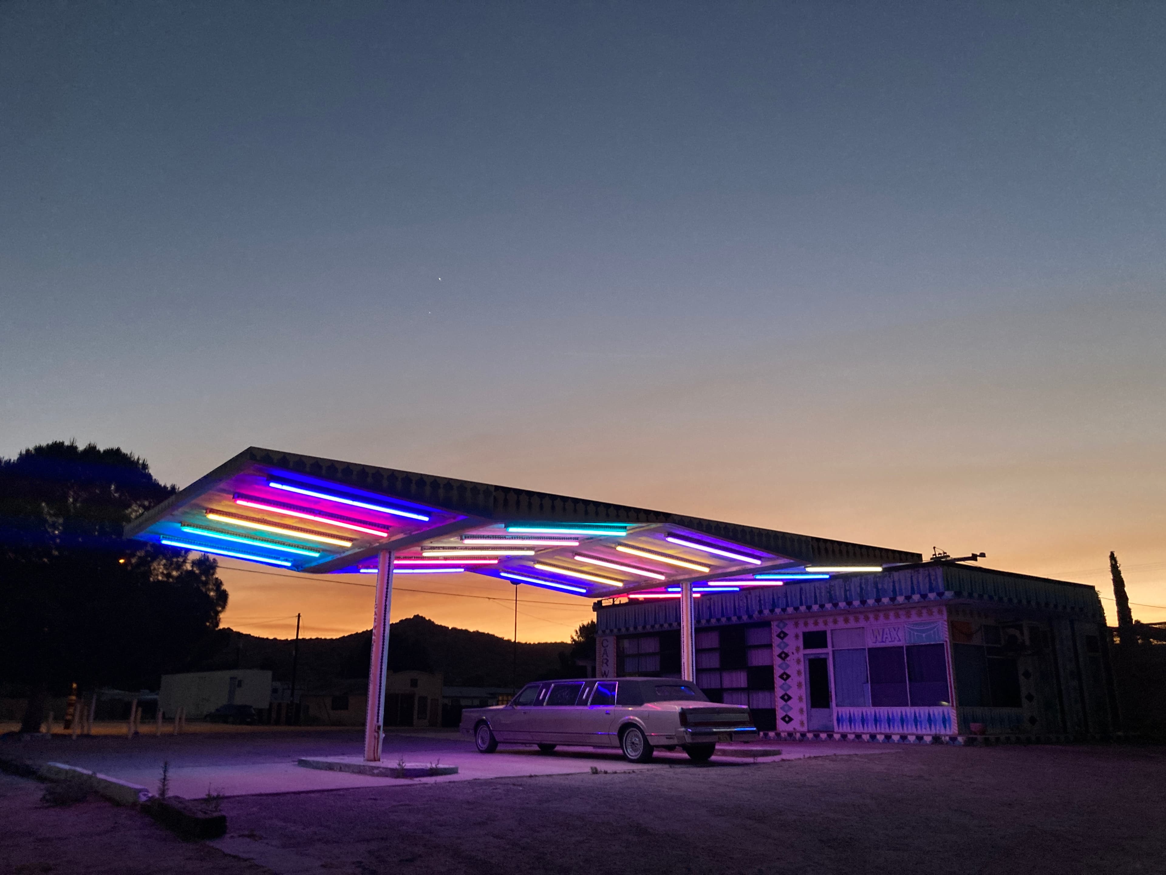 A neon-lit gas station canopy at dusk with a vintage limousine parked underneath